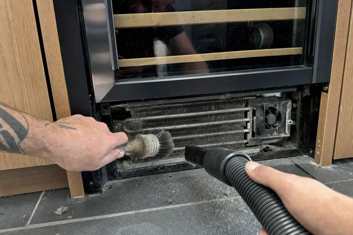Brush and vacuum hose clearing a dust-matted condenser coil behind the lower grille of a wine cooler
