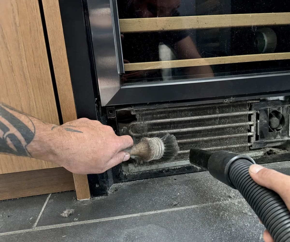 Condenser coil being brushed clean behind the grille of a built-in wine cooler