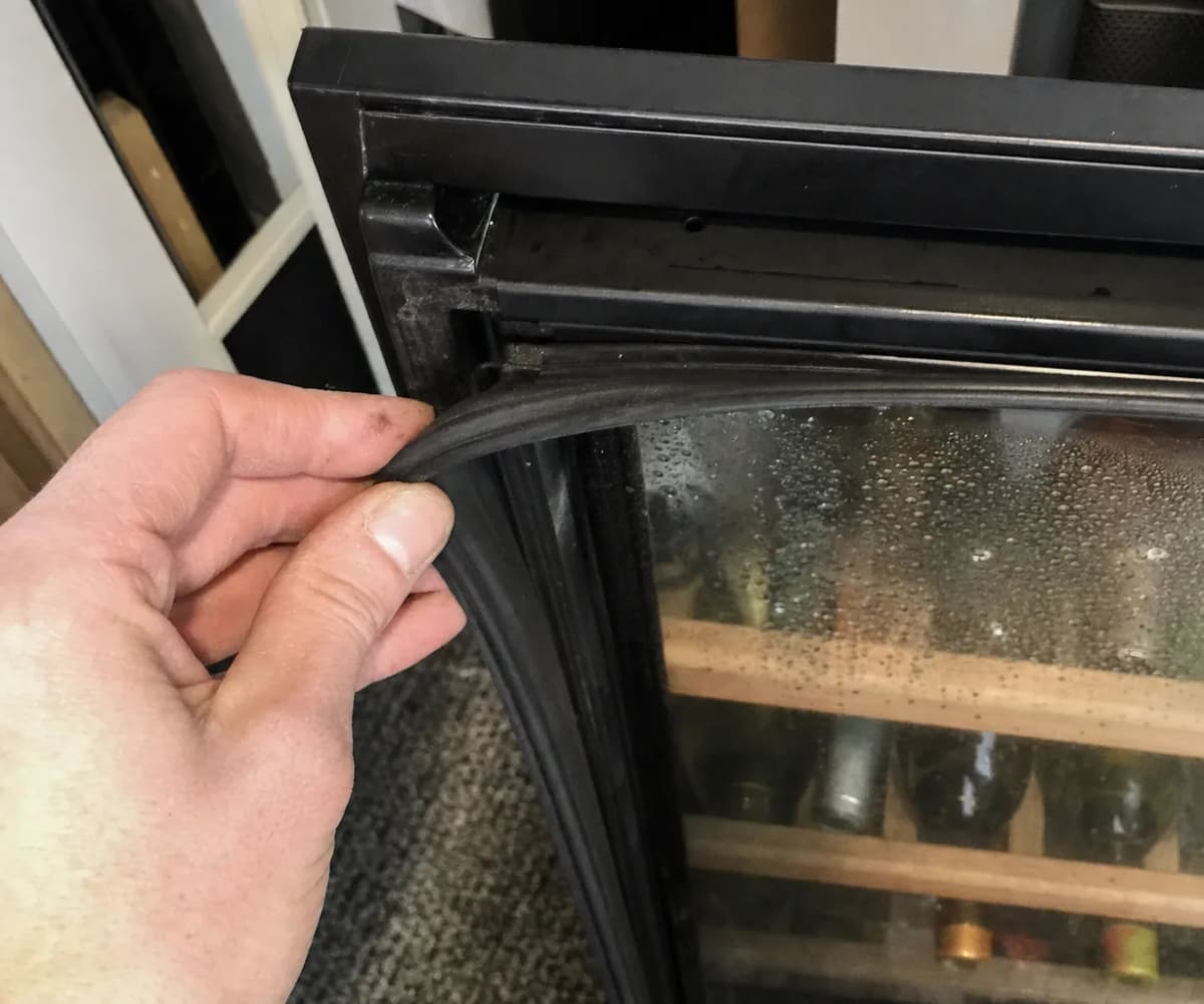 Close view of a hand lifting the door gasket at the corner, condensation beaded on the glass behind
