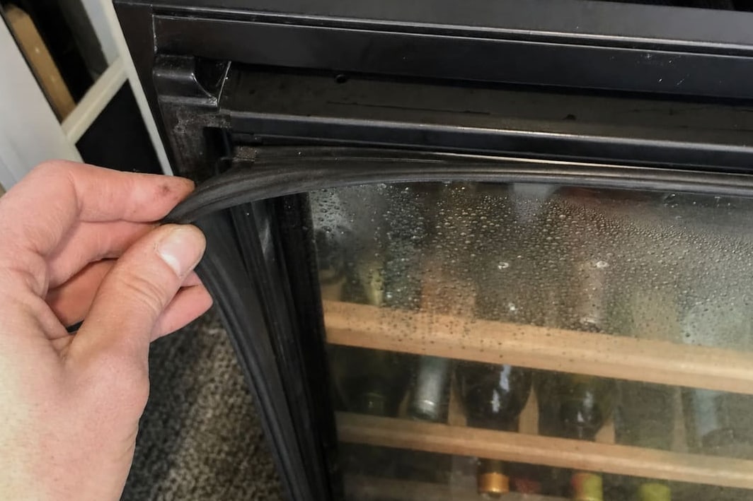 Hand pulling the magnetic door gasket away from the frame of a wine cooler, condensation on the glass behind