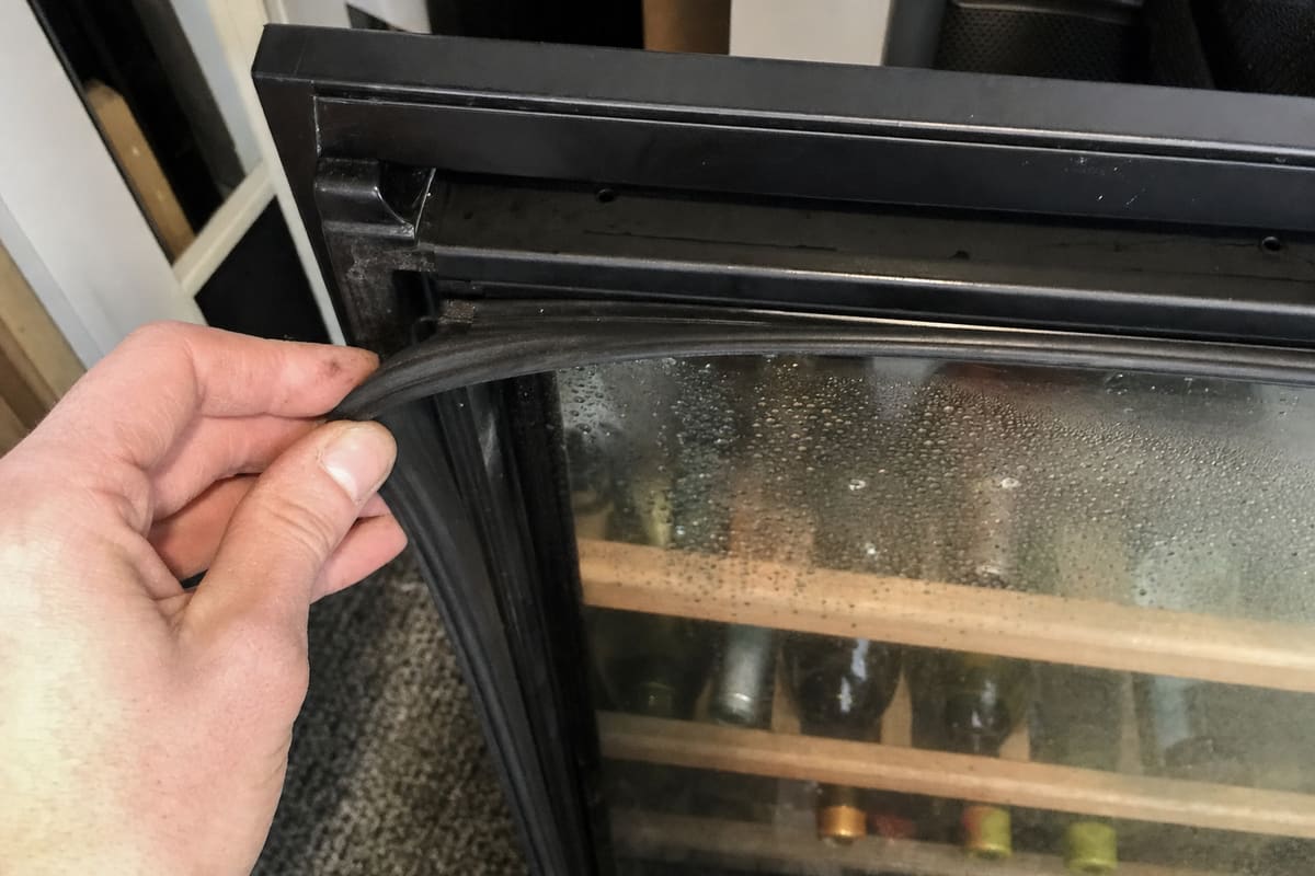Hand pulling the magnetic gasket away from the frame of a wine cooler door, glass fogged behind