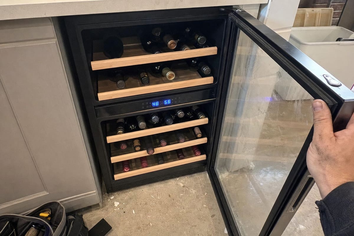 Under-counter dual-zone wine cooler with the glass door open, bottles on wooden shelves and a blue LED display between the zones