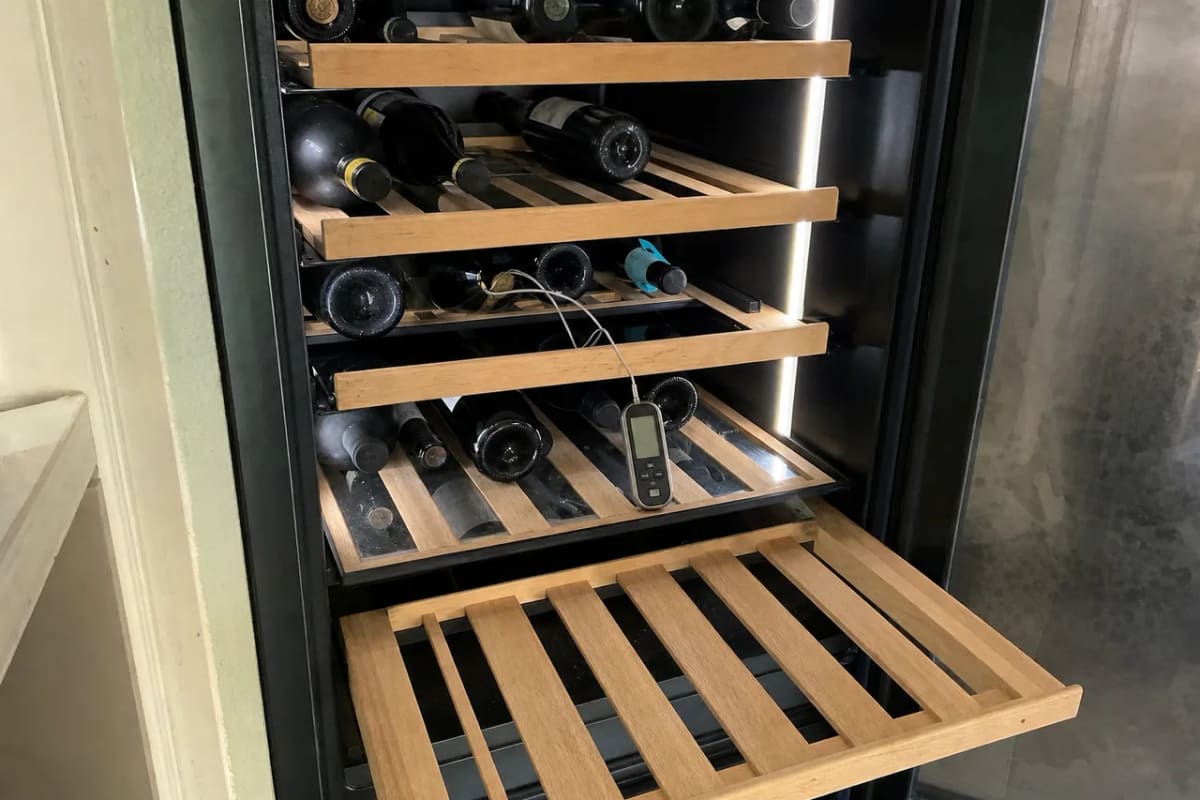 Bottles on wooden shelves inside a wine cooler with a probe thermometer reading the shelf temperature