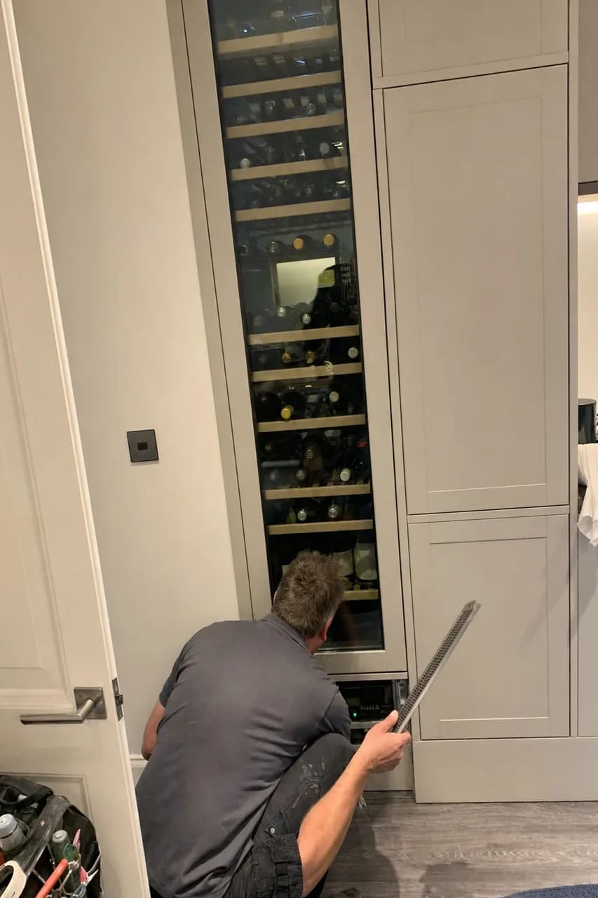 Technician working at the lower grille of a tall built-in wine column loaded with bottles