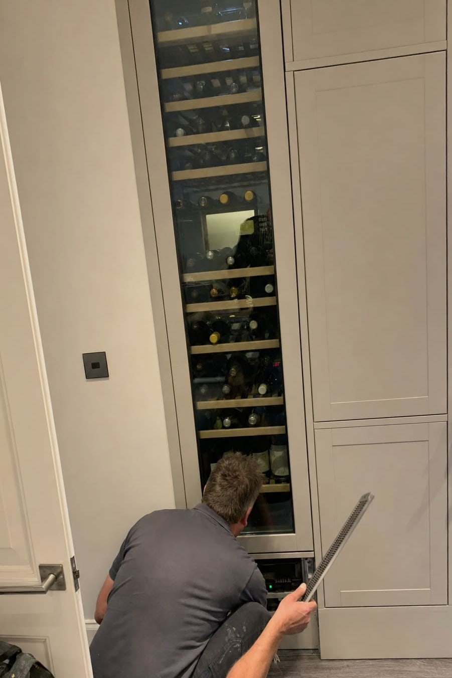 Technician crouched at the base of a full-height built-in wine column, working at the lower grille