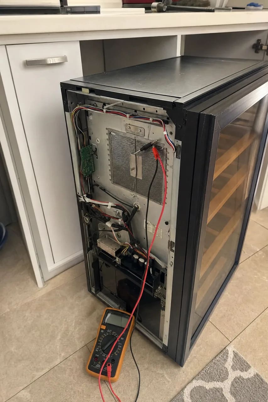 Back panel removed from a thermoelectric wine cooler, heat sink and control board exposed, multimeter connected