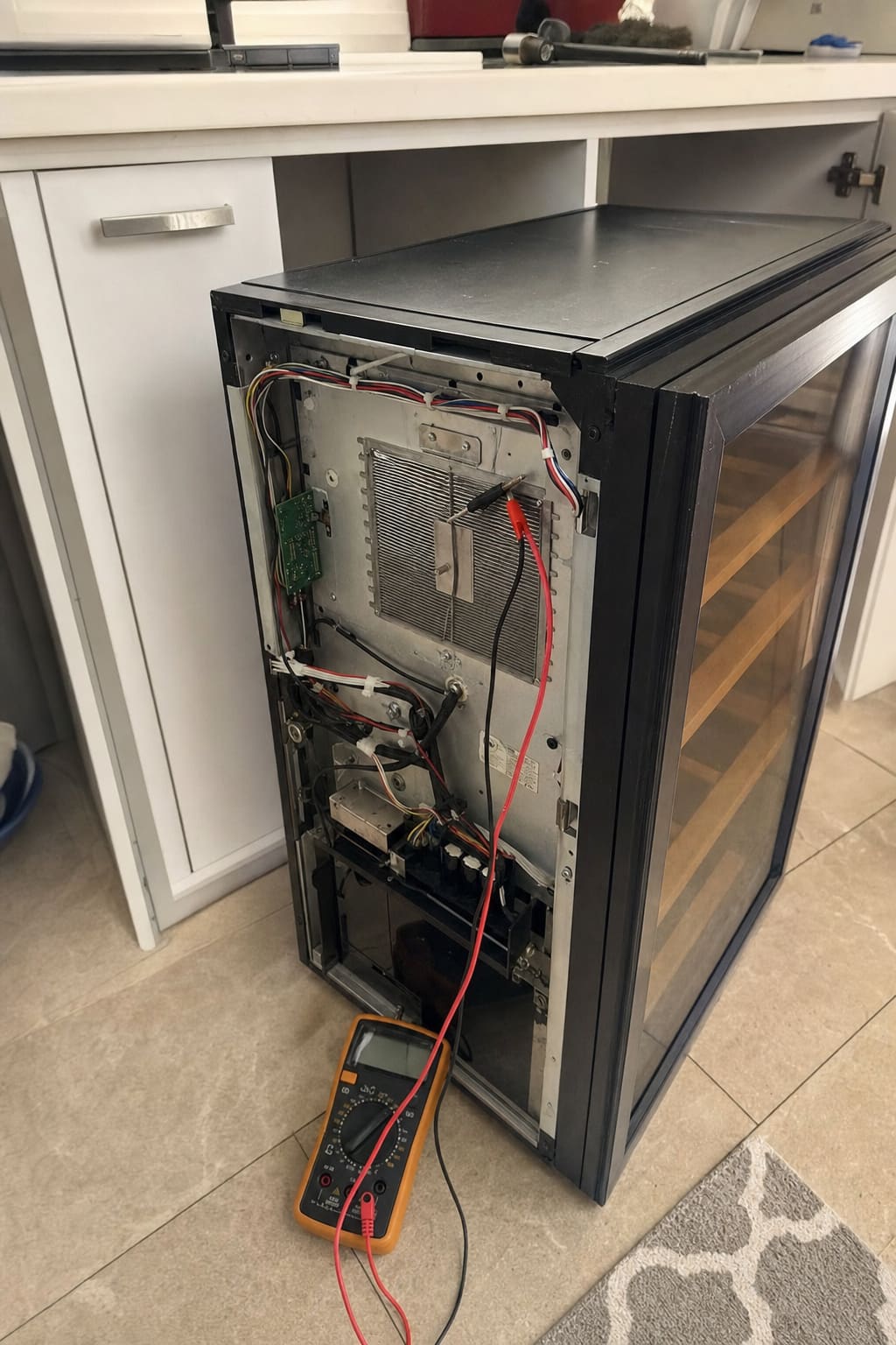 Back panel off a thermoelectric wine cooler, heat sink and control board exposed with a multimeter on the floor