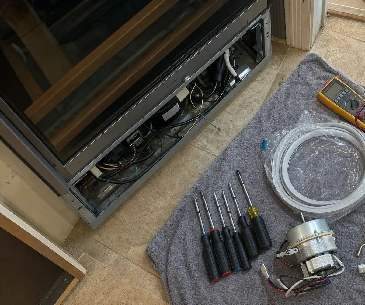 Screwdrivers, a multimeter, a replacement fan motor and tubing beside an open wine cooler service bay