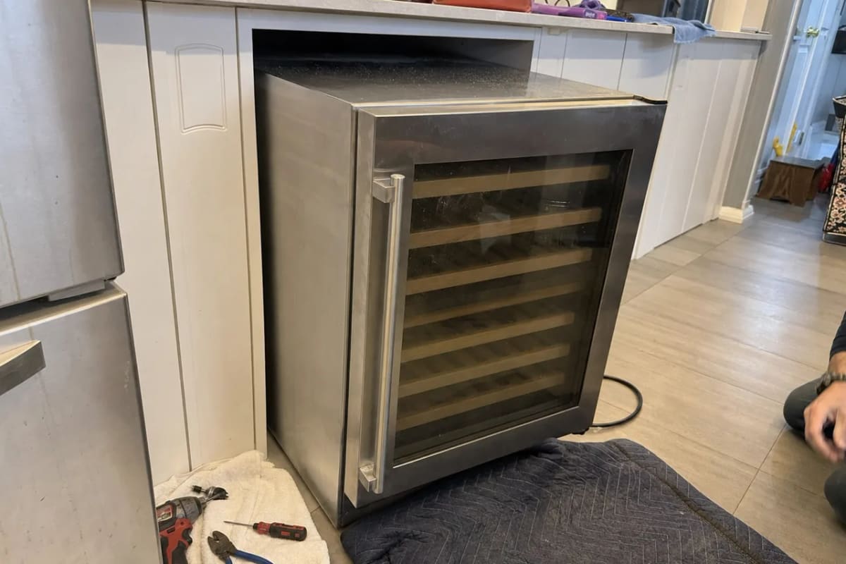Stainless under-counter wine cooler pulled forward out of its cabinet opening, power cord looped behind, tools on the floor