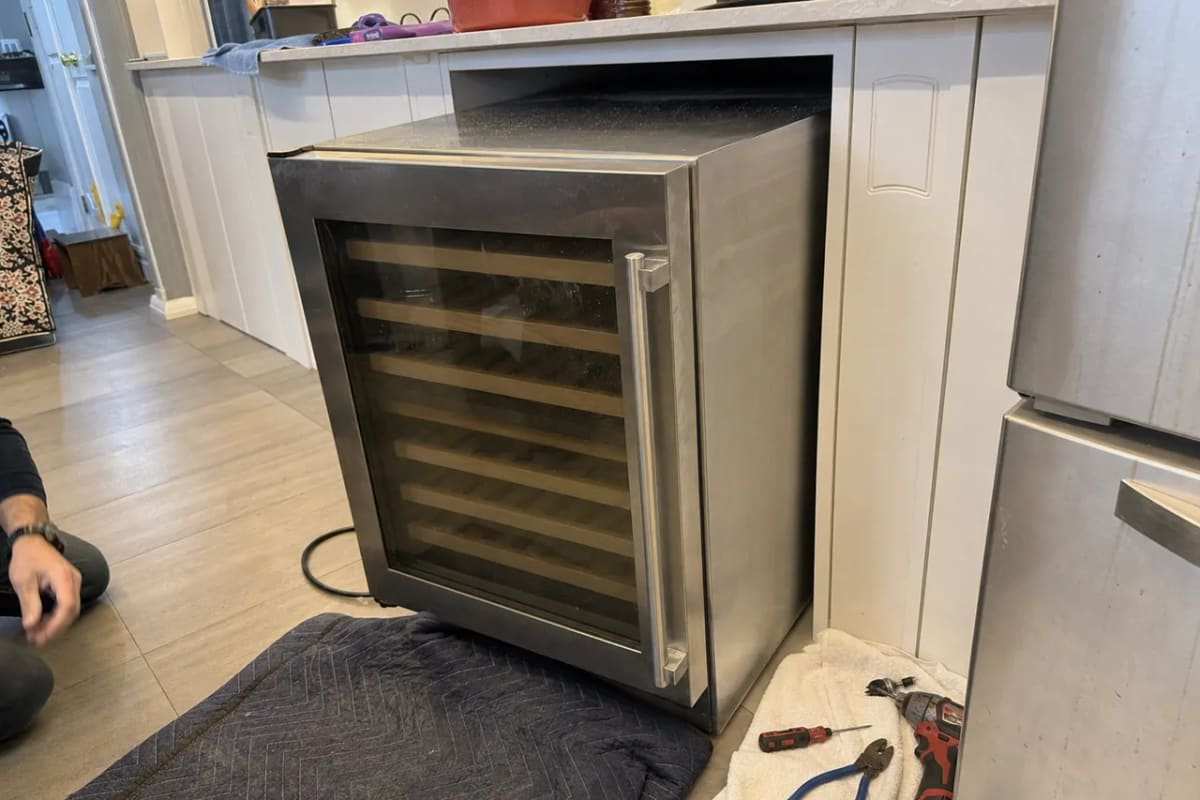 Under-counter wine cooler drawn out of a narrow kitchen cabinet opening, tools laid out beside it