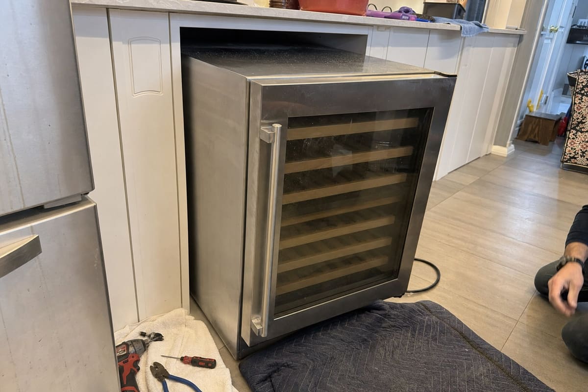 Stainless under-counter wine cooler pulled clear of its cabinet opening for service, tools on the floor